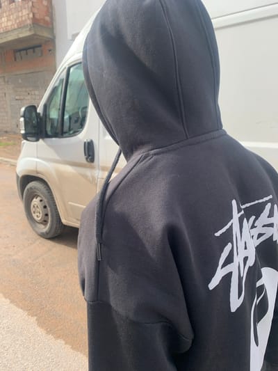 The image depicts a close-up, over-the-shoulder view of an individual from behind, located in Ameur عامر, Morocco. The subject is wearing a black hooded sweatshirt with the hood drawn up, obscuring their head. A prominent white "Stussy" logo is visible on the upper right rear portion of the sweatshirt. The hood's black drawstring, tipped with a dark aglet, is visible on the right side.
In the background, positioned to the left of the subject, is a dirty white commercial van, possibly a Fiat Ducato or similar model, parked or idling on a yellowish-brown unpaved or dusty road surface. The van's front driver's side wheel with a grey hubcap, mud-splashed lower body, side mirror, and partial windows are visible. Beyond the van, in the upper left background, is a structure constructed from exposed red bricks and grey concrete, appearing to be unfinished or under construction. The scene is illuminated by natural daylight. No discernible actions or interactions are occurring.