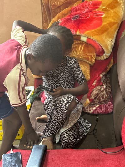 In Gidan Waziri, Nigeria, two children are seated on a couch covered with blankets. The child in the foreground, wearing a patterned dress, holds a smartphone. Another child, wearing a red and white jacket, leans over the first child, looking at the phone. The surrounding area is dimly lit, with a red blanket and a brown couch visible. A power bank and a charging cable are also present.