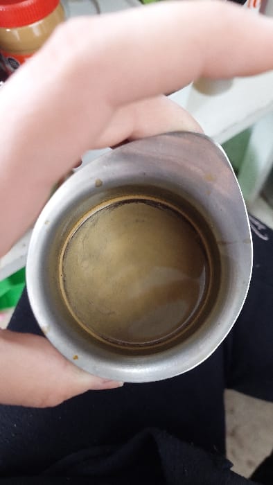 An overhead photograph displays a light-skinned hand gripping a silver-colored metal pitcher. The pitcher contains a shallow quantity of light brown liquid, consistent with coffee or espresso, topped with a visible crema layer. Minor dark residue is present along the interior rim of the pitcher. The hand's fingers are wrapped around the upper portion of the pitcher. In the blurry background, a red-lidded jar containing a light brown substance is partially visible to the upper left, adjacent to a light-colored shelf or surface. Dark fabric, likely part of the individual's attire, is visible in the lower foreground. The scene is situated in Tlaxcala City, Mexico.