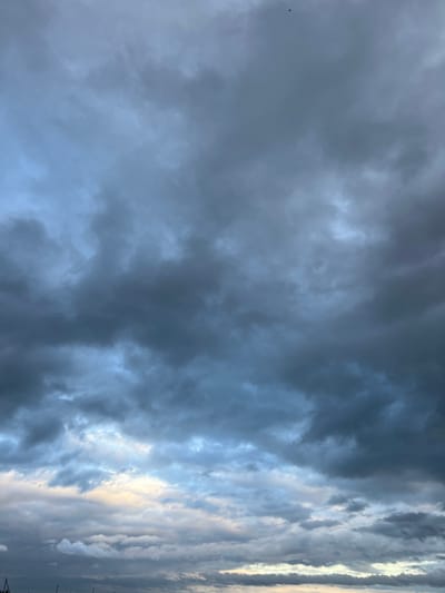 The image shows a dramatic sky filled with dark, heavy clouds, suggesting an impending storm. The clouds are dense and textured, with varying shades of gray and blue, hinting at atmospheric depth. In the far upper center of the image, a tiny, dark silhouette, possibly a bird or a distant aircraft, is visible against the overcast sky. The lower portion of the image reveals a sliver of lighter sky, with hints of yellow and pale blue, suggesting the presence of the sun attempting to break through or the approaching dawn or dusk. The scene is devoid of any specific landmarks or elements that would tie it to Калодзежскі сельскі Савет, Belarus, or any other urban location. The focus is entirely on the atmospheric conditions of the sky.
