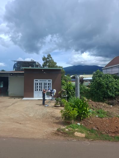 The image depicts an outdoor scene in Thôn 2, Vietnam, under a cloudy sky. In the foreground, a paved road is visible. The mid-ground features a man standing on a concrete surface, tending to laundry on a drying rack. To his right, there are various plants and a pile of dirt. Behind the man is a brown building with a white door and windows. To the left of this building is a larger structure with a large, closed garage door. In the background, there are more buildings and lush green vegetation, with a mountain range visible in the distance. The overall impression is of a residential or commercial area with elements of domestic activity.