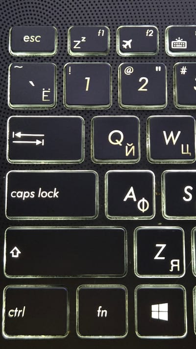 This is a close-up, high-angle shot of a black laptop keyboard. The keys are illuminated from beneath with a white light, creating a glow around their edges. The keys have both Latin and Cyrillic characters on them. Visible keys include "esc", "f1", "f2", "~", "Ё", "!", "1", "@", "2", "#", "3", a symbol resembling two arrows pointing away from each other, "Q", "Й", "W", "Ц", "caps lock", "A", "Ф", "S", a large blank key with an upward arrow symbol, "Z", "Я", "ctrl", "fn", and a Windows logo key. The background is a dark, textured surface with a dotted pattern, likely the palm rest of the laptop. The image provides no information about the city of Lebedyan, Russia, or any other specific location.