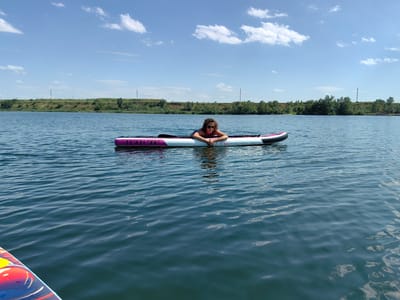 A woman is lying prone on a pink and white stand-up paddleboard on a calm body of water. She is wearing sunglasses and a dark-colored bikini top. The paddleboard has "11'X31"X6\"" printed on its side. The background features a low, grassy embankment with scattered trees and power lines, under a blue sky with scattered white clouds. The image appears to be taken on a sunny day. The location is identified as the city of Samoilov, Russia.