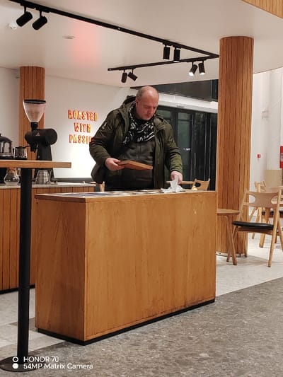 An individual in an olive jacket and patterned scarf stands behind a wooden counter. He holds a wooden object in one hand and a white cloth in the other. A coffee grinder sits on a table to the left. In the background, "ROASTED WITH PASSION" is illuminated on a wall. Wooden pillars and furniture fill the space, with light fixtures overhead. The floor is a speckled grey. A camera watermark is at the bottom left.