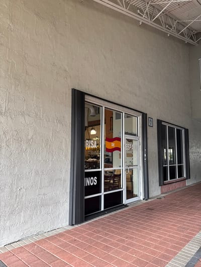 A building facade is visible, featuring two windows. The lower window has a sign indicating "España" and "RISA...NOS," and displays the Spanish flag. Reflected in the glass are interior elements, possibly tables and chairs. A smaller window is situated above. The building's exterior has a textured, light-colored surface. A white metal structure is to the left and a brick walkway to the right of the facade.