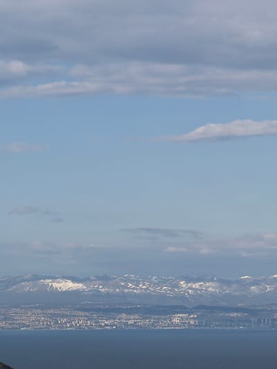 The image shows a view of Zagorje, Croatia from an elevated perspective. The sky is predominantly blue with scattered clouds. In the distance, a mountain range is visible, partially covered with snow. The city stretches along the coastline, with buildings and structures densely packed together. The sea is dark and calm. The overall scene depicts a landscape blending urban and natural elements.