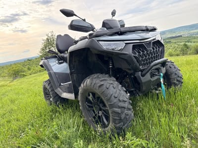 The image shows a dark gray all-terrain vehicle (ATV) parked on a grassy hill. The ATV is facing forward, angled slightly to the right. The front wheels are prominent, showcasing their large, knobby tires. The front of the ATV features a honeycomb-patterned grill and a protective bar. A turquoise strap hangs down from the front. The background consists of a green hillside with scattered trees and a distant view of a village or town. The sky is overcast with light gray clouds. The scene appears to be set in a rural or semi-rural location. While the exact location within Blagoevo, Bulgaria is not directly discernible from the image, the landscape features suggest a setting in the outskirts or surrounding areas of the city.