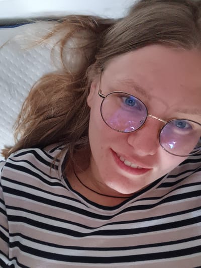 The image is a close-up, high-angle shot of a young woman with light brown hair and blue eyes, wearing round, gold-rimmed glasses and a black, beige, and white horizontally striped shirt. She is lying down on a white, textured surface, possibly a mattress. Her head is tilted back, and she is looking up towards the camera with a slight smile. The lighting in the image suggests an indoor setting, with reflections of light visible on her glasses. There is no visible information in the image that indicates a specific location in the city of Koledino, Russia.