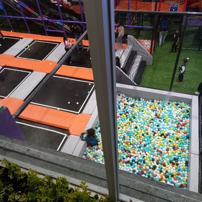 The image depicts an indoor recreational facility located in Tumbaco, Ecuador, observed from an elevated viewpoint through a window or glass barrier. Green foliage is visible outside the structure in the bottom left foreground.
The facility is divided into multiple zones. On the left, a large trampoline area features several interconnected rectangular trampolines with black jumping surfaces bordered by orange and gray safety padding. Small, multi-colored balls (yellow, white, green) are scattered across some trampolines. Approximately five individuals are present in this area; one person in a white top and dark pants stands on a trampoline in the mid-left, while two others in dark and light tops are visible further back.
Adjacent to the trampoline area, in the lower-right quadrant, is a large, rectangular ball pit with gray edges, densely filled with plastic balls in shades of white, light blue, yellow, red, purple, and black. A person wearing a blue jacket is partially submerged in the ball pit, leaning against its edge.
To the right of the ball pit and trampolines, an area covered with artificial green turf is visible, enclosed by black safety netting. A gray set of stairs leads up to an elevated platform within this section. Several individuals are on the turf, including one person in black and red clothing standing in the mid-right. In total, approximately 10-12 individuals are engaged in various activities across the facility. Overhead, structural elements with what appear to be colored lights are visible in the top left. The environment indicates an active entertainment center.