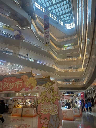 The image depicts the interior of a multi-story shopping mall in Licheng District, China. The scene is dominated by a large, brightly lit atrium extending upwards through multiple levels, each featuring curvilinear balconies adorned with strings of golden decorative lights. A prominent glass skylight forms the ceiling of the atrium, admitting natural light.

On the ground floor, a temporary market structure is set up, featuring a large archway with the Chinese characters "FA FA FA 发财好市" (Prosperity Market) and "IN CITY 济南" (Jinan). The archway is decorated with traditional Chinese art, including images of fireworks, a cartoon pig/boar figure, and a person alongside a tree with the English text "8 HORSES." Beneath this archway, several vendor stalls with light-colored awnings are arranged, displaying various merchandise such as jewelry and crafts.

Approximately 10-15 individuals are visible on the ground floor, engaged in walking, browsing market stalls, and observing their surroundings. A few individuals are also visible on upper levels.

Hanging vertically from a higher level, a large banner advertises "活活鱼 要现选" (Live fish, choose on site) and "吃200 送200" (Spend 200 get 200), indicating a promotional offer. The banner also notes "L5," likely referring to the fifth floor. On a higher floor balcony to the left, a large, purple inflatable object shaped like a hand holding a yellow stick with "bang!" written on it is visible. Escalators are partially visible on several levels. The overall environment is well-lit, combining natural light from the skylight with extensive artificial illumination.