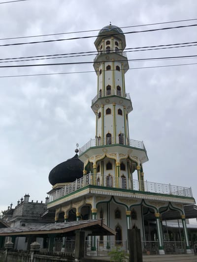 The image displays a tall, ornate minaret, which is part of a mosque, set against an overcast sky. The minaret is primarily white with distinctive green and gold decorative patterns, featuring multiple levels with small windows and balconies enclosed by metal railings. A small, blue-tiled dome or cupola tops the minaret. To the right of the minaret, a large, dark-colored mosque dome is visible. Several dark power lines crisscross the upper left and central portions of the frame, partially obscuring the minaret.
The sky is cloudy and grey, indicating an overcast day. No people or specific activities are depicted; the scene is a static architectural view. The overall aesthetic of the mosque, with its minaret and dome, is consistent with Islamic architecture found in Indonesia. The specific location provided is Bale, Indonesia. There is no visible text in the image.