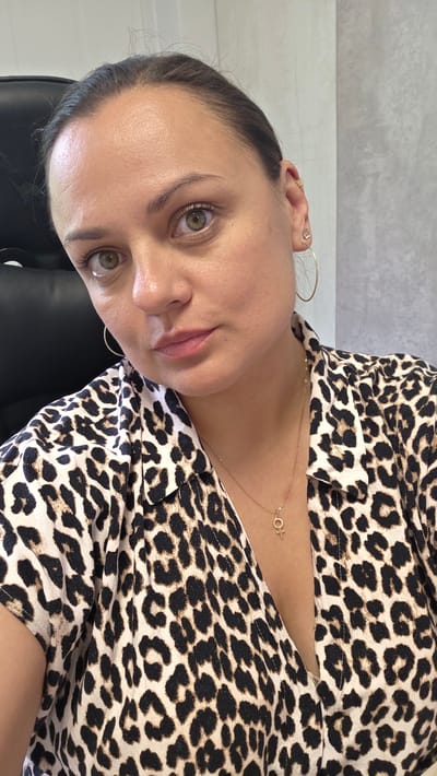 This is a portrait of a young woman taken from a slightly high angle. She has light brown hair pulled back from her face and is wearing a leopard-print blouse with a V-neckline. A delicate gold necklace with a small circular pendant is visible on her chest. She has large, expressive eyes with light green irises. Her lips are painted a soft pink. She is wearing hoop earrings in one ear, and a small stud earring in the other. The background appears to be indoors, with a hint of a dark surface behind her head and a textured light gray wall. The lighting suggests it is daytime. The overall impression is one of a casual, personal photograph. There is no visible text in the image.