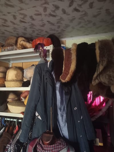 The image depicts the interior of a storage space in посёлок 1-го отделения совхоза "Масловский", Russia. The area seems to be filled with various clothing items, fabrics, and accessories stacked on shelves and racks. Several fur hats and garments with fur trim are visible. There is also a dark coat on a hanger. Mannequin heads, some wrapped in plastic, are placed on shelves. Additional textiles and clothing items are piled beneath the coat and racks. The background wall has a patterned covering. Overall, the storage space appears cluttered with a diverse array of clothing and fashion-related items.