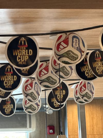The image shows a ceiling decorated with circular McDonald's "FIFA World Cup Meal" signs and images of Adidas soccer balls. The McDonald's logo is visible on some of the signs. The environment appears to be indoors, possibly a McDonald's restaurant, with a wooden ceiling and part of a window or opening in the lower left corner. The scene is devoid of people or any actions being performed. The image is taken from a low angle looking upwards at the ceiling decorations. The specific location of Port Coquitlam, Canada is not visually inferable from the image itself.