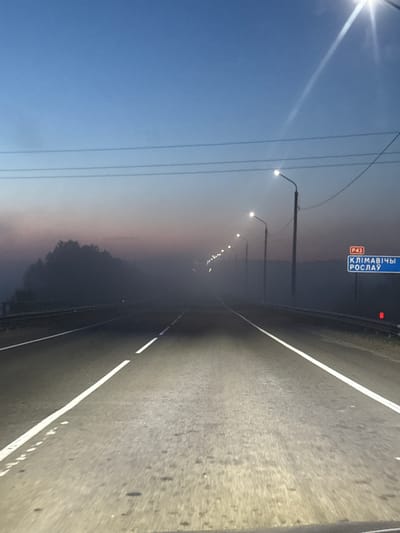 The image shows a road in Krychau, Belarus. The sign on the left reads "Klimavichy Roslavl" (the cities), road P43. The road is paved with white lines marking the lanes. Street lights illuminate the road. The sky is blue near the top of the image, transitioning to a gradient of pink/orange near the horizon. Power lines are visible on the left side of the image. There are trees on the left side of the road. The light suggests it is dawn or dusk.