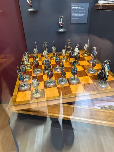 Here is a description of the image:

The image shows a chess set with figures dressed as 18th-century Austrian soldiers on display in a glass case. The board is made of light and dark wood squares, and the figures stand on silver and gold circular bases. A sign next to the chess set identifies it as "Spielfiguren des Schachspiels" (Chess figures) by Georg Ignatius Baur, a Biberach native (1727-1790). The location is likely a museum or historical exhibit in Biberach an der Riß, Germany.