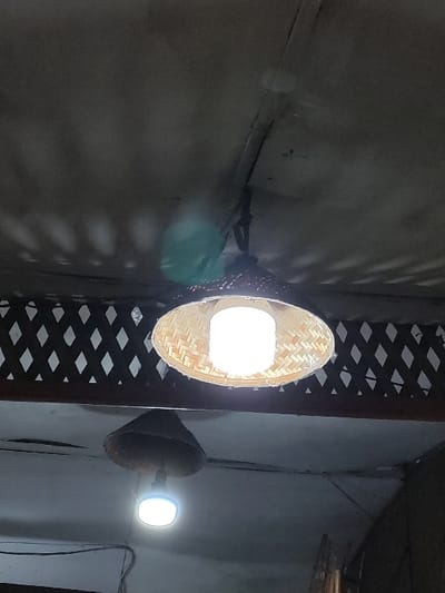 The image displays an interior ceiling scene with two artificial light sources. The primary subject is a circular, woven light-colored bamboo or rattan lampshade, centrally located and mounted to a light-grey ceiling. A bright white LED bulb is visible within this shade, emitting light downwards and casting a distinct diamond-patterned shadow onto the ceiling surface directly above. Below and to the left of the main fixture, a second, darker, conical or bell-shaped lampshade is partially visible, also containing a white LED bulb that emits light downwards. A dark brown wooden lattice structure with a diamond pattern is present in the upper background, behind the main light fixture, suggesting a decorative partition or architectural element. The ceiling shows a seam or crack running diagonally across the frame. Lens flare is evident from the brighter light source. No people, discernible actions, or interactions are visible. The specific location within Staufenberg, Germany, cannot be determined from the provided visual information.