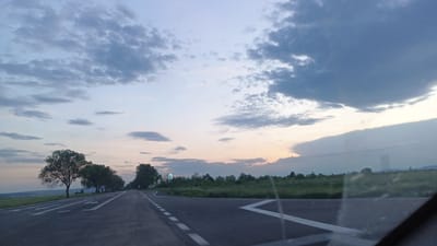 The image depicts a two-lane asphalt road in a rural area, likely near Sokolovo, Bulgaria. The road has white painted lane markings and is oriented toward the horizon. Trees line the left side of the road, casting long shadows. To the right of the road, a grassy field extends towards the horizon. The sky is filled with clouds, ranging from light gray to dark blue, suggesting either early morning or late evening light. The photograph appears to have been taken from inside a vehicle, indicated by the presence of the windshield's edge in the lower right corner. There are no people visible in the image.