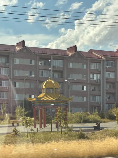 This media file, captured in Дорожный, Russia, presents an outdoor scene dominated by a multi-story brick apartment building. The building has a reddish-brown roof and numerous windows, with some air conditioning units visible on the exterior. In front of the building, a bright yellow, fish-shaped structure, possibly a playground element or public art, stands out. Several red posts are also present, forming part of what could be a fence or seating area.
To the left of the building, power lines stretch across a bright blue sky filled with fluffy white clouds, suggesting it is daytime with pleasant weather. A thin, metallic structure, possibly an antenna, is visible on the roof of the building. To the right of the building and the yellow structure, a paved pathway runs alongside a strip of dry, yellowish grass and scattered green trees and bushes. The overall impression is of a residential area on a clear day. There is no visible text in the image.
