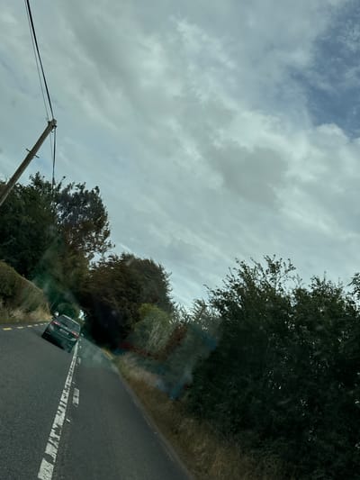 A dark-colored car is driving on a paved road, turning to the left. The car is in the middle of the road, with a dashed white line on its right and a solid yellow line on its left. The road is lined with trees and grass on both sides. The sky is cloudy. There is a utility pole on the left side of the road, with wires running across the sky. The scene appears to be in a rural area of New Ross Municipal District, Ireland, given the road markings and vegetation.