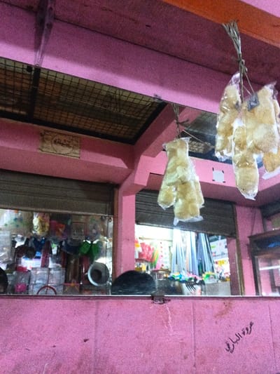 The image depicts an indoor market or commercial area located in Kidapawan, Philippines, characterized by a dominant faded pink color on its structural elements. An overhead dark brown beam spans the upper portion of the frame. Below this, multiple pink-painted concrete beams and columns form the internal structure. A metal mesh grate is affixed to the underside of a pink beam in the upper left quadrant.
Two distinct clusters of light-colored items, individually packaged in clear plastic bags and resembling puffed snacks or crackers, are suspended by twine from the overhead dark brown beam in the upper right.
Through visible openings, interiors of adjacent retail spaces are discernible. The shop on the left features a partially lowered gray roll-down security shutter above a display area containing various household goods, including clear plastic containers, kitchenware, and other items. The shop on the right is brightly lit from within, showcasing numerous vertically arranged objects, possibly pipes or fabric rolls, along with other colorful merchandise.
A light-colored, rectangular sign with dark, illegible script is mounted on a pink beam in the upper left. On the lower right, dark cursive script, consistent with Arabic characters reading "عروة البارئ", is inscribed on the pink wall or counter surface. No individuals are present or engaged in any depicted activity.