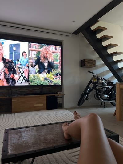 The image depicts an indoor scene, likely a living room or a common area in a home, with elements suggesting a modern and somewhat eclectic design. The dominant objects are a staircase with dark wooden treads and risers, a vintage motorcycle parked against a white wall, and a large television screen displaying a scene from a movie or TV show.
In the foreground, a person's legs and feet are visible, resting on a textured rug next to a dark-colored coffee table. The person has dark nail polish on their toes. The television screen is the focal point for many of the scenes it displays: the top portion shows a woman with long, curly blonde hair in an outdoor setting with greenery and a brick building; the middle section shows a woman in a light blue top and a patterned skirt sitting on a folding chair; and the bottom section shows a person holding a small, fluffy black dog.
The motorcycle appears to be a classic model, parked at an angle. Beside it, on the wall, is a wooden crate labeled with text that is partially visible, including "BEVERAGES" and "BEVERAGE." The overall lighting suggests it is daytime, though the specific time of day is not discernible. The setting appears to be indoors, possibly in a well-lit room with natural light coming from an unseen source or from the open spaces of the house. The presence of the television, furniture, and decorative items like the wooden crate suggest a lived-in, comfortable space. The scene conveys a relaxed atmosphere, with someone enjoying entertainment or a break. The location context indicates this is in Mandelieu-la-Napoule, France.