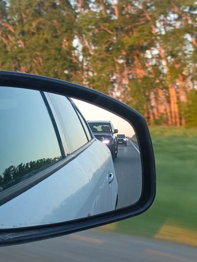 The image shows a side mirror of a white car. The mirror reflects the road behind the car, with several vehicles following. A forest is visible in the background. The location is likely in a rural area of the Ural region of Russia, as suggested by the presence of birch trees and the name of the city. The scene is from the perspective of the driver of the white car.