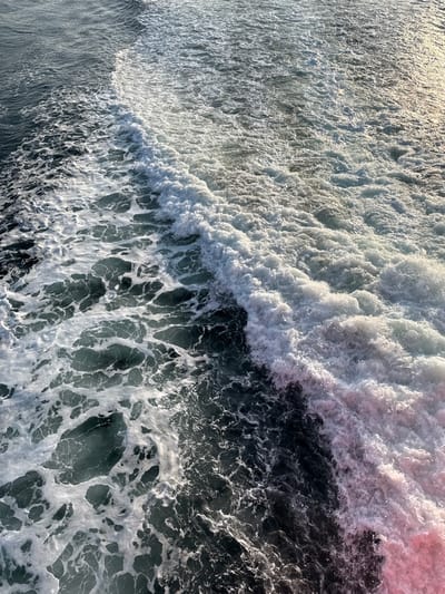 The scene depicts turbulent water, likely a vessel's wake, located in the waters near Palaiopoli, Greece. A prominent diagonal crest of white, frothy foam extends from the upper left to the lower right of the frame. To the left of this crest, the water is a darker blue-green with dispersed patches of white foam. To the right, the water is extensively churned into dense white foam and brighter areas reflecting sunlight, with a subtle pinkish hue visible in the foam towards the lower right. No individuals or specific objects other than the water itself are visible. The interaction of water and implied motion creates dynamic patterns of foam and ripples under bright environmental illumination.