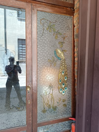 The image shows a double door, with one door being clear glass and the other being frosted glass with a peacock design. The door is set in what appears to be the city of Mezzana Sant'Antonio, Italy, evidenced by the architecture visible through the glass. A person is reflected in the clear glass door, holding a smartphone and taking a picture. A fire extinguisher is visible near the base of the door with the peacock design, which is positioned near a brick wall and a dark brown doorframe. The overall style is likely Art Nouveau or a similar artistic period.
