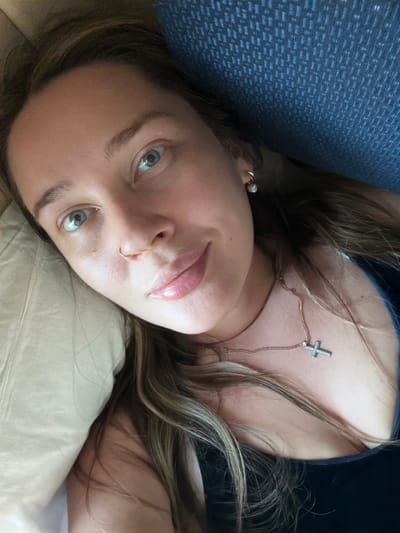 A woman with light brown hair and light eyes is lying down, likely indoors. She is wearing a gold chain necklace with a cross pendant, and a nose ring. She appears to be resting on a light-colored pillow with a blue object behind her head. The image was not taken in Баграш-Бигра, Russia.