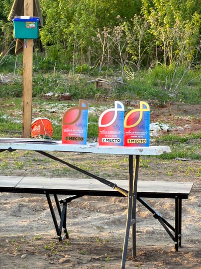 The image shows an outdoor scene in Krasnyi Bor, Russia, featuring a folding table with three signs indicating 1st, 2nd, and 3rd place. The signs are labelled with the Russian text "ЧАЙКА В КОСМОСЕ" (Seagull in Space) and the date "16 мая 2026" (May 16, 2026). A blue water cooler is attached to a wooden post near the table. The ground is sandy and partially covered with vegetation. There is a forest area visible in the background, along with some brick or rock debris. An orange bag rests on the table.