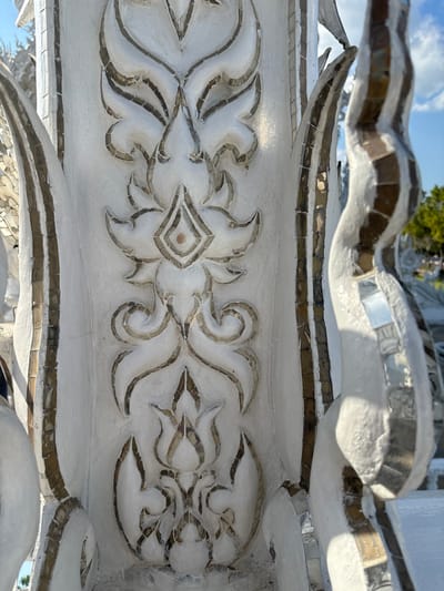 A detailed, vertical image displays an architectural element of the Wat Rong Khun (White Temple) located in Ban Rong Khun, Thailand. The structure is composed of white stucco or plaster, featuring a repeating pattern of traditional Thai floral or flame motifs rendered in high relief along a central vertical panel. These designs are precisely outlined and partially filled with embedded fragments of mirrored glass, creating a mosaic effect. To the left and right of the central panel, curved architectural forms, similarly constructed from white material and accented with vertical strips of mirror mosaic, are partially visible. The scene is illuminated by direct sunlight, causing reflections from the mirrored surfaces. A clear blue sky with distant, out-of-focus green foliage is observable in the upper right background, indicating an outdoor setting. No human figures or discernible activities are present.