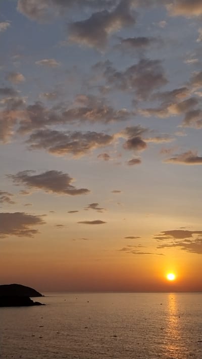 The image depicts a sunset over the ocean, with a silhouette of a headland on the left side of the frame. The sky is filled with scattered clouds, ranging in color from pale blue and white to shades of pink and orange, indicating the setting sun. The sun itself is a bright, glowing orb positioned on the right side of the horizon, casting a shimmering reflection on the calm water. The ocean surface is relatively smooth, with small dark shapes that appear to be boats or buoys scattered across it. The headland on the left is dark and forms a distinct shape against the colorful sky. The overall scene suggests a peaceful and scenic coastal environment. The location is identified as Tuy Phong 4, Vietnam.