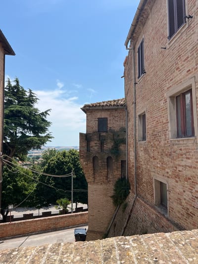 This image captures a view of an ancient Italian town with old brick buildings and lush greenery. The foreground is dominated by a weathered stone wall, suggesting a vantage point from an elevated position. To the right, a brick building with dark-shuttered windows and a tiled roof stands prominently. A lower brick wall with a tiled coping extends along the side of this building, forming a parapet.
Further into the scene, an ornate brick tower with arched openings and a darker tiled roof juts out from another building. Vines cling to the brickwork of this tower. To the left, a tall evergreen tree partially obscures the view of other brick buildings and more greenery.
Below, a road winds through the town, with a pedestrian crossing visible in the distance. A black trash bin is placed beside the road. Beyond the immediate buildings and road, a rolling landscape of hills, dotted with trees and some structures, stretches towards the horizon under a bright blue sky with scattered white clouds and contrails. The architecture and the style of the landscape strongly suggest a location in Italy. Given the context of Barbara, Italy, this scene likely depicts a street or viewpoint within the historic center of the town, showcasing its medieval architecture and scenic surroundings.
