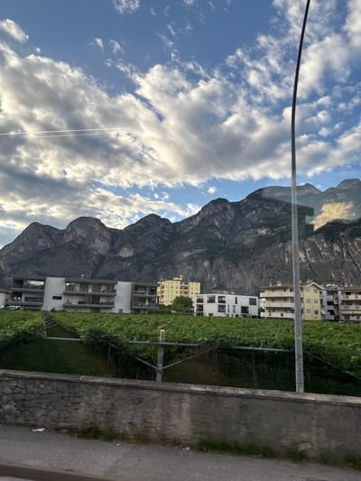 The image captures a scenic view of Mezzolombardo, Italy, dominated by a majestic mountain range in the background. The sky above is a canvas of blue adorned with dramatic clouds, with a faint contrail suggesting the passage of an aircraft. In the mid-ground, a modern apartment building with a white facade and multiple balconies stands out, bordered by lush green vineyards. Adjacent to it, a yellow apartment building adds a splash of color to the scene. A lamppost stands tall in the foreground, partially obscuring the view of a stone wall and a paved road below. The overall impression is one of a tranquil, picturesque town nestled against a breathtaking natural backdrop.