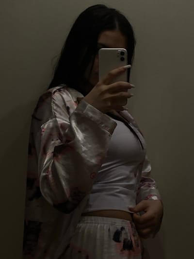 A single individual, gender appearing female, is depicted indoors against a plain, light-colored wall in Vakhitovo, Russia. The person has long, dark hair partially obscuring their face and is wearing a light-colored fitted top underneath an open, long-sleeved satin robe and matching pajama bottoms. The robe and bottoms feature a floral or abstract pattern in shades of pink and dark hues. The individual's left hand, adorned with long, light-colored nails, holds a silver-colored smartphone with a dual-lens camera, positioned as if taking a photograph. The right hand is partially visible, tugging at the waistband of the pajama bottoms. The scene is dimly lit with indirect lighting.