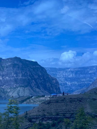 The image depicts a wide shot of a mountainous landscape with a body of water in the foreground. The sky is a deep blue with scattered clouds. A prominent, dark mountain range dominates the left side of the frame, with a lighter, more distant mountain range visible in the background. The terrain in the mid-ground appears to be terraced and arid, with several small buildings clustered on a hillside overlooking the water. The water itself is a vibrant blue. The overall impression is one of a remote and rugged natural setting. The location is Shamilkala, Russia.