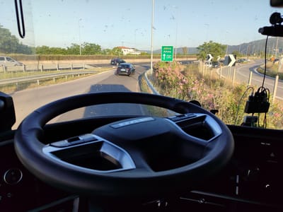 The image provides a first-person view from inside a vehicle, likely a truck or bus given the "DAF" logo prominently displayed on the steering wheel, looking out through a windshield marked with dust or debris. The steering wheel dominates the lower center of the frame, and parts of the dashboard are visible, including a screen showing "Mon 1 Jun" and "Android Auto."

The scene outside is a bright, sunny day with clear skies. The vehicle is on a highway approaching an exit. In the mid-ground, a dark-colored car is on an exit ramp, followed by a dark grey SUV. A green road sign, central to the image, clearly displays "Αθήνα Athina" with an upward arrow and the motorway designation "A1," indicating the direction to Athens. This, along with the provided location context, identifies the setting as the Municipal Unit of Atalanti, Greece, likely along the A1 motorway.

The highway is bordered by guardrails and lush vegetation, including numerous trees, green bushes, and clusters of pink flowering plants, adding a vibrant touch to the roadside. Streetlights are visible along the highway. In the background, distant mountains rise under the bright sky. To the right of the main exit, a winding road with chevron signs indicating curves is visible, with several parked cars. A silver van can be seen on the main highway to the left, indicating ongoing traffic. The image captures an active road environment under strong daylight conditions.
