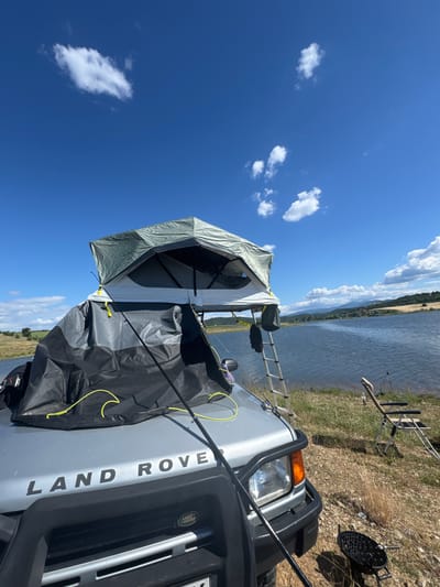 The image depicts an outdoor scene near the city of Drenkovo, Bulgaria, featuring a silver Land Rover Discovery with a roof tent. The vehicle is parked on a grassy, dry terrain next to a large body of water. The roof tent is partially deployed, with a ladder leaning against the side of the vehicle for access. A black cover is draped over the front of the vehicle, extending to the roof tent. Fishing rods are visible near the water's edge, with a folding chair set up for fishing. A small charcoal grill is also present on the ground. The sky is blue with scattered white clouds, and a distant hilly landscape forms the horizon. The scene suggests a recreational activity, likely camping and fishing, in a natural setting.