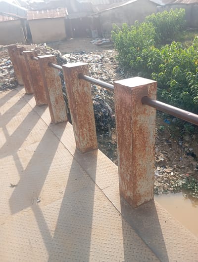 The image shows a rusty railing made of square metal posts and a dark metal pipe along the top, casting long shadows on a metal grate floor. Behind the railing, a scene of disarray unfolds with piles of garbage and debris. In the background, several dilapidated structures with corrugated metal roofs suggest a densely populated or informal settlement. To the right, a dense growth of green bushes borders the garbage-strewn ground, and a murky body of water is visible. The overall impression is one of neglect and poor sanitation in an urban or semi-urban environment. This scene is located in Gwagwa, Nigeria.