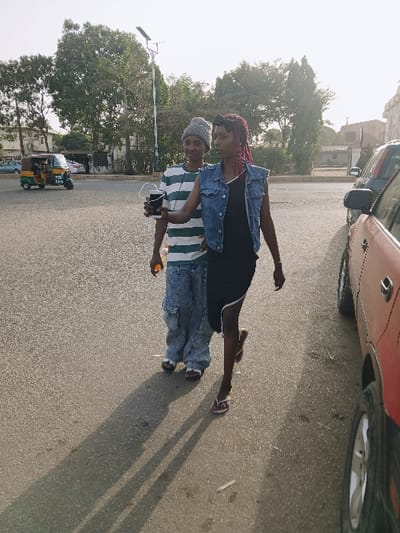 The scene is set outdoors on an asphalt surface in the city of Kungulu, Zambia, near Kungulu School, under bright daylight creating long, distinct shadows, suggesting either morning or late afternoon. Two females are depicted in motion, walking towards the viewer's right.
The individual on the left, positioned slightly behind, wears a grey knit beanie, a green and white horizontally striped long-sleeved shirt, baggy light blue cargo-style jeans, and light-colored sandals. She holds a small orange object in her right hand and looks towards the camera.
The individual on the right, in the foreground, has reddish-pink braided hair adorned with a black ribbon/band. She wears a black sleeveless dress featuring a white-trimmed diagonal slit on the right thigh, over which she wears a blue denim sleeveless vest. She wears light-colored flip-flops. Her left hand is extended forward, holding a black cup with white wired earbuds dangling from it. Her right leg is raised, indicating a walking stride.
In the background, a yellow and green auto-rickshaw (tuk-tuk) is visible on a road on the far left. A partial view of a red vehicle is seen on the far right, with only its front fender, wheel, and part of the door visible. A tall white street light pole, equipped with a solar panel, stands behind the individuals. Various trees and distant buildings complete the background environment.