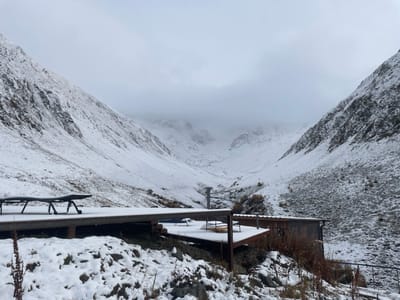 This image depicts a snow-covered mountain valley in Juta, Georgia, under an overcast sky. In the foreground, a multi-tiered wooden deck extends from the right, partially covered with snow. Two dark-colored lounge chairs, also dusted with snow, are positioned on the upper left portion of the deck. Adjacent to the deck on the right is a single-story, dark-wood building with a dark roof, partially obscured by snow-covered terrain and sparse dry vegetation. Two light-colored cylindrical metal structures, possibly vents or chimneys, protrude near the deck. The background features steep, snow-laden mountain slopes flanking both sides of the valley, which recedes into a thick layer of low-hanging clouds. Exposed dark rock and dry, autumnal vegetation are visible amidst the snow on the slopes. No individuals or active movements are present in the scene.