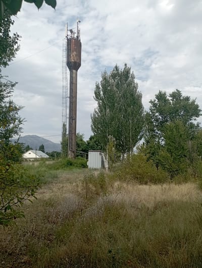 The image depicts an outdoor scene with a prominent, weathered water tower topped with telecommunication antennas. The tower is made of concrete at the base and a rusty, cylindrical metal structure at the top. A ladder runs along the side of the concrete base. To the right of the tower, a white, corrugated metal container or small building is visible.
In the background, a white house with a gabled roof and a small window is situated amidst vegetation. Behind the house, a mountain range with a rugged, rocky slope rises against a cloudy sky. The foreground and mid-ground are filled with tall, dry grass and scattered green bushes and trees. Several trees, including a tall, slender one with lighter-colored leaves, are visible. The overall environment suggests a rural or semi-rural setting. The location is identified as Vostochnoe, Kyrgyzstan. There are no people visible in the image.