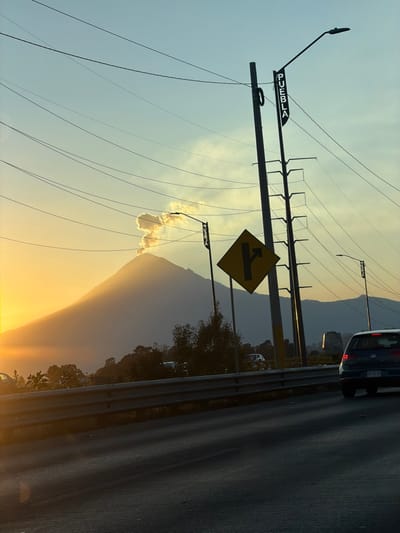 The image is taken on a road near Puebla, Mexico. A diamond-shaped road sign indicates a left turn ahead. Overhead electrical lines traverse the sky, attached to metal poles. A light blue sky fades into a yellow sunset over a mountain. A passing dark car is visible on the road. The scene has a warm, golden hue due to the setting sun.