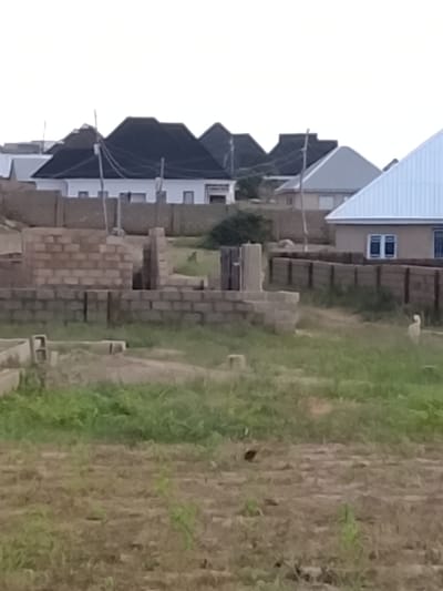 This is a daytime, medium-angle exterior shot of an under-construction residential area. The foreground consists of dry, patchy grass and dirt. In the middle ground, a structure is being built with concrete blocks, showing foundations and partial walls. To the right, a wooden fence separates this construction site from a completed residential area. The completed houses feature modern architecture with dark, pitched roofs and light-colored walls, some with visible windows. One house has a light blue, corrugated metal roof. A solitary, light-colored dog stands near the wooden fence in the middle ground. The background is dominated by a hazy sky, suggesting an overcast or early morning/late evening setting. The location is identified as Barakin Acha, Nigeria.