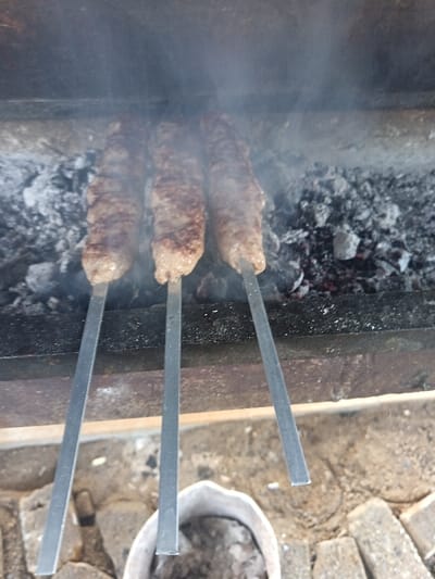 Three elongated meat items, skewered on flat metallic rods, are being cooked over glowing charcoal and ash within a rectangular metal barbecue grill. White smoke emanates from the embers, partially obscuring the upper portion of the scene. The meat appears partially cooked with visible browning. The lower ends of the skewers extend downwards towards an outdoor ground surface comprised of dirt and irregularly shaped light-colored paving stones. A light-colored, open-mouthed container, possibly ceramic or metal, is positioned on the ground in the lower-right foreground. The scene is located outdoors in Kasri Arifan, Uzbekistan.