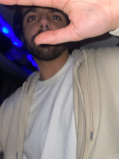 A man's face with a beard and dark hair is partially visible. His left hand is in the foreground, obscuring part of his face. He wears a white t-shirt under a beige hooded sweatshirt. Blue lights are blurred in the background. The image is slightly rotated, tilting the subject's face to the left.