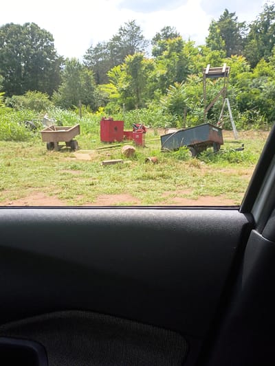 The image, taken from inside a vehicle, looks out onto a rural landscape in Hartman, United States. Through the car window, various pieces of farm equipment are scattered across a grassy field. These include a rusted metal cart, a red tractor component, and a wooden wagon. In the background, lush green trees fill the horizon under a partly cloudy sky. The foreground features the interior of the vehicle, specifically the door panel with dark textured materials.
