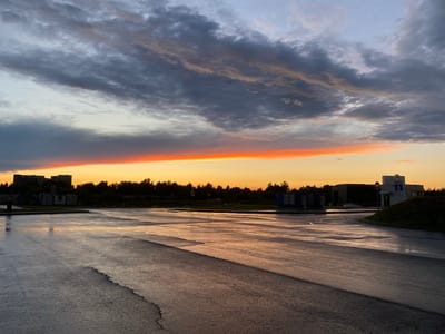 This is a horizontal, eye-level, medium shot photograph taken at sunset in the city of Bratovshchina, Russia. The foreground is dominated by a wet asphalt parking lot or road, reflecting the sky's colors. The asphalt has visible cracks and is uneven. In the middle ground, there is a tree line with some structures visible, including modular buildings, a darker building with multiple windows, and a modern-looking building with signage and a rounded hill to its right. A white car is parked in front of the darker building. The sky is filled with dramatic clouds, with a bright orange band of light at the horizon where the sun is setting, transitioning to darker gray and blue clouds higher up. The overall mood is serene and atmospheric.