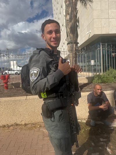 An outdoor scene in Lod, Israel, depicts two males near the Lod District Court building under a partly cloudy sky. In the foreground, a young adult male, smiling at the camera, stands equipped in an Israel Police tactical uniform consisting of a grey long-sleeved shirt, matching cargo pants, a black tactical vest displaying an Israel Police emblem patch on the left sleeve, and a black backpack. He carries a black assault rifle with a magazine and optics, held vertically. A neon yellow band is visible around his waist. To his right, slightly behind and lower, a bald adult male in a dark t-shirt and pants squats on the paved ground, looking towards the left with a slight smile. The background features the distinctive light-colored, multi-story facade of the Lod District Court building, fronted by a dark metal fence and a security camera. A prominent palm tree trunk stands between the primary subject and the building. To the far left, a red fire hydrant and white parked vehicles are visible behind a separate metal fence. The immediate ground is paved with red interlocking bricks.